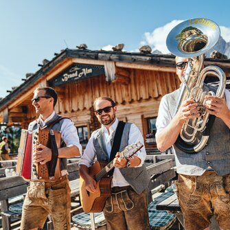 Three musicians in traditional attire with accordion, guitar and tuba in front of Brand Alm hut in sunny Ramsau am Dachstein | © Christine Höflehner
