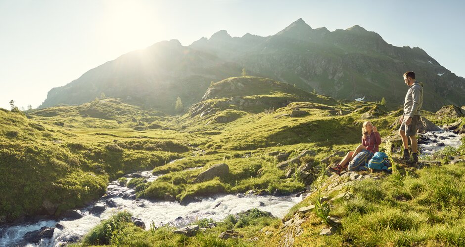 Two hikers rest by a mountain stream in the sun with green alpine pastures and mountain peaks in the background | © Peter Burgstaller