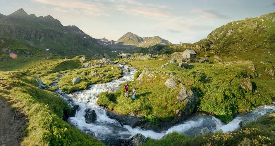 Two hikers by a clear mountain stream in lush alpine scenery with huts and mountain peaks in the background | © Peter Burgstaller