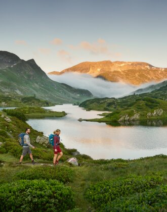 Two hikers walk along Lake Giglach, surrounded by green mountains, morning sun and mist drifting through the valley | © Peter Burgstaller