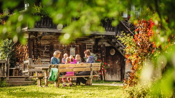 Family with children sits on wooden benches in front of rustic hut, surrounded by flowers, trees and summer light | © photo-austria Höflehner