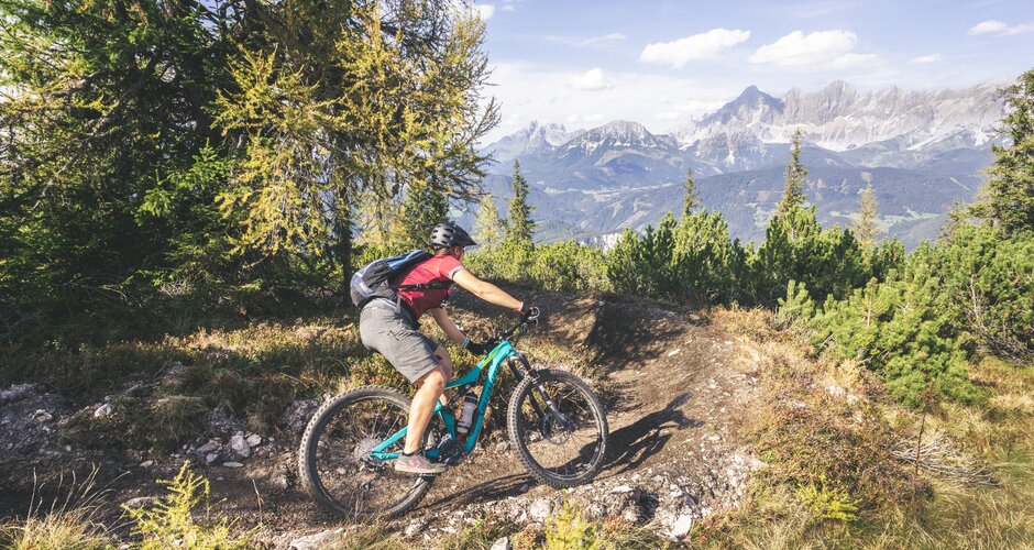 Mountain biker on a narrow forest trail at Reiteralm with the Dachstein massif in the background under a blue sky | © Georg Knaus