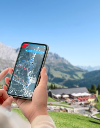 Person holds smartphone with interactive trail map, background shows gondola, huts and mountain panorama in Ski amadé.