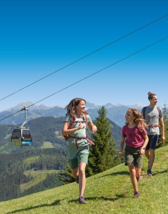 Group of four hikers enjoys a sunny day on the alpine meadow with gondola view and wooden hut in the background in Ski amadé.