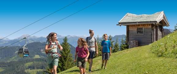 Group of four hikers enjoys a sunny day on the alpine meadow with gondola view and wooden hut in the background in Ski amadé.