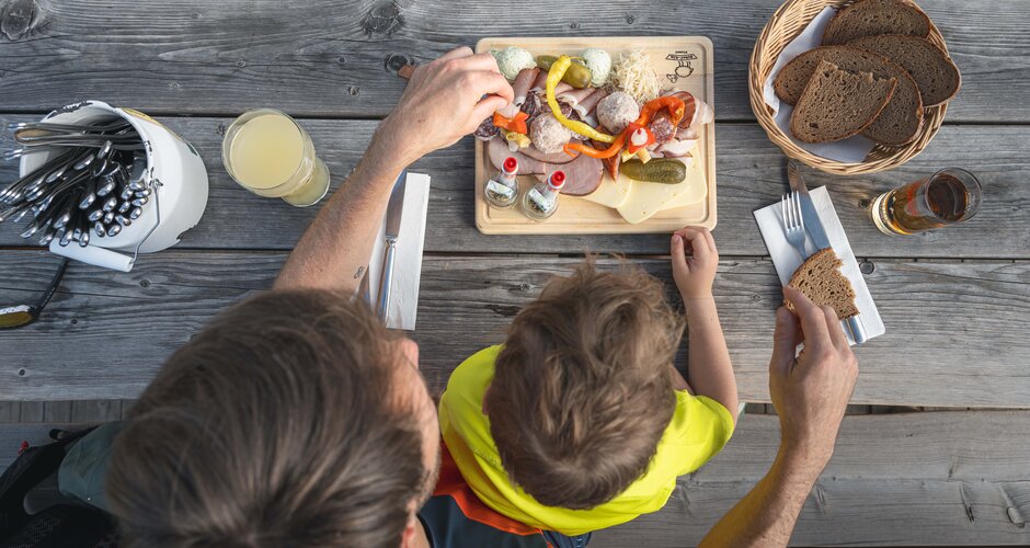 Father and child enjoy cold cuts, cheese and bread at a wooden table, seen from above at a rustic alpine hut.