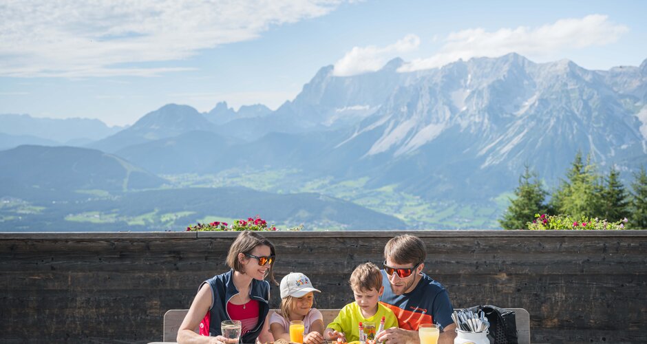Family with two kids enjoys snack board and drinks at a wooden table on a summer hut terrace with scenic view of the Alps.
