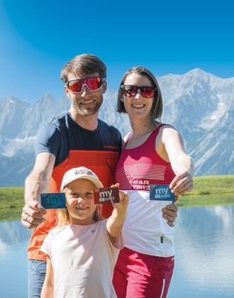 Smiling family holds up their my Ski amadé cards at a mountain lake with a stunning Alpine backdrop behind them.