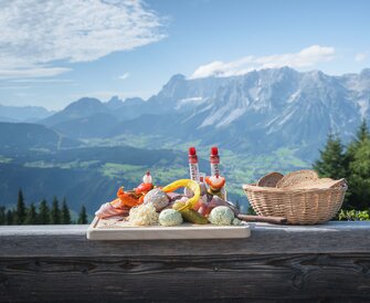 Traditional snack board with cheese, sausage, spreads and bread on a bench with a stunning Alpine panorama behind.