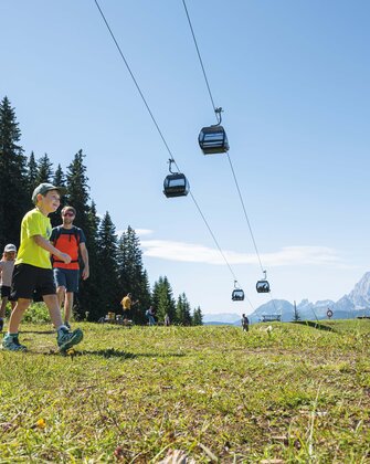 Eine Familie wandert bei Sonnenschein auf einer Almwiese unter schwebenden Gondeln mit Blick auf die Berge von Ski amadé.