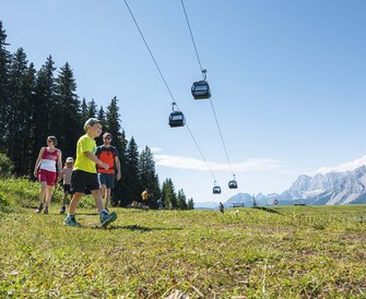 A family hikes across an alpine meadow beneath gondolas, enjoying the sunshine and mountain views in the Ski amadé region.