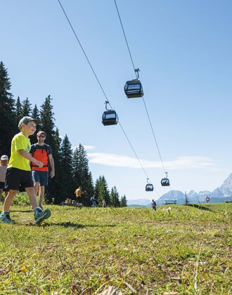 Eine Familie wandert bei Sonnenschein auf einer Almwiese unter schwebenden Gondeln mit Blick auf die Berge von Ski amadé.