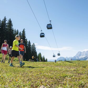 A family hikes across an alpine meadow beneath gondolas, enjoying the sunshine and mountain views in the Ski amadé region.