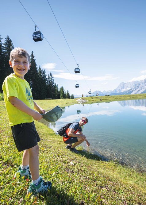 A smiling boy plays at the edge of a clear mountain lake while his father kneels behind him, gondolas float above the scenery.