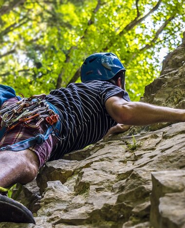 A climber with helmet and full gear ascends a steep rock face surrounded by forest, bathed in warm summer light.