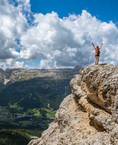 A hiker with backpack stands with arms raised on a rocky summit plateau overlooking a wide green alpine valley.