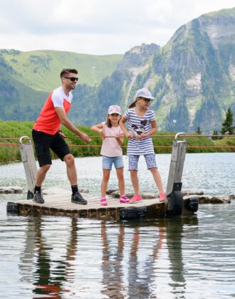 Man and two girls on a raft pulled by rope across a mountain lake, surrounded by green alpine scenery and hills.