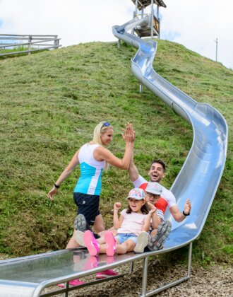 Two kids and their parents laugh while sliding down a long metal slide in the mountains – a joyful alpine family moment.