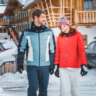 A young couple dressed in winter clothes strolls hand in hand through the snowy ski resort, smiling happily at each other. | © Ski amadé