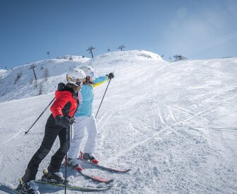 Two best ager female skiers stand on a snowy slope, smiling and looking ahead, with ski lifts in the background.