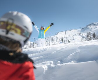 A skier (best ager) in blue jacket with yellow sleeve waves happily in the snow, with snowy slopes in the background.