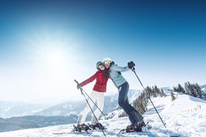 Two female skiers hug on the slope, wearing ski goggles and helmets, enjoying the Alpine panorama in the background.