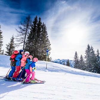 Six children in colorful ski outfits ski closely together down the slope, smiling and enjoying the snow.