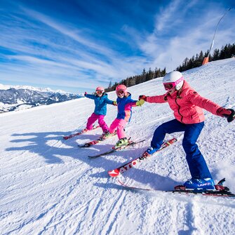 Drei Kinder fahren mit bunten Skianzügen nebeneinander auf der Piste, halten sich an den Händen und lachen im Schnee.