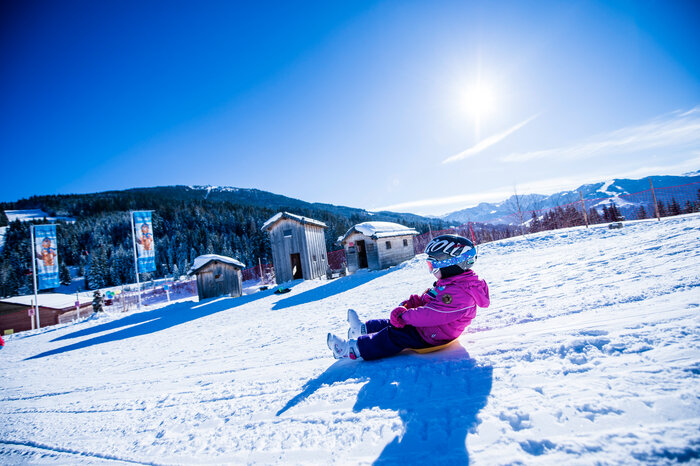 Kind mit Helm und Skibrille fährt lachend mit dem Schlitten eine verschneite Piste hinab, Holzhütten dahinter.