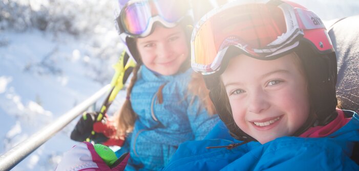 Two kids in colorful ski suits and helmets sit smiling on the ski lift, enjoying the sun and snowy landscape.