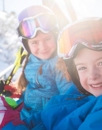 Two kids in colorful ski suits and helmets sit smiling on the ski lift, enjoying the sun and snowy landscape.