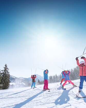 Kinder in bunter Skikleidung heben Skistöcke freudig in die Luft, vor verschneiten Bergen und Pisten.