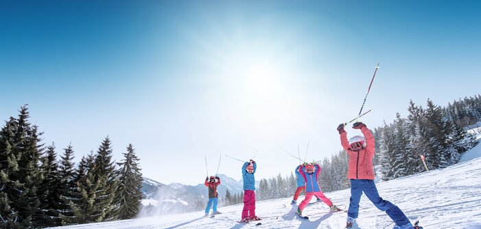 Kids in colorful ski outfits raise ski poles happily in the air, with snowy mountains and slopes in the background.