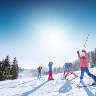 Kinder in bunter Skikleidung heben Skistöcke freudig in die Luft, vor verschneiten Bergen und Pisten.
