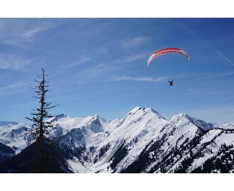 Ein Gleitschirmflieger schwebt hoch über den schneebedeckten Bergen von Großarl unter blauem Himmel.
