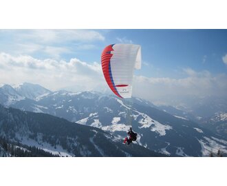 Paragleiter mit rot-blau-weißem  Schirm fliegt über von Wald und schneereichen Pisten bedeckte Berge