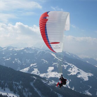 Paraglider with red, blue and white glider flies over mountains covered in forest and snowy pistes