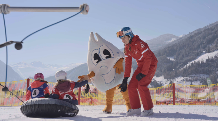 Zwei Kinder sitzen auf Reifen im Schneepark Dorfgastein, Maskottchen Gasti und Skilehrerin begrüßen sie mit Lächeln | © Dorfgasteiner Bergbahnen