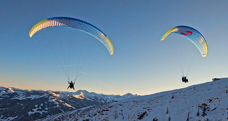 Zwei Personen gleiten mit Gleitschirmen im Tandemflug über verschneite Berglandschaft bei Sonnenuntergang in Dorfgastein | © Dorfgasteiner Bergbahnen