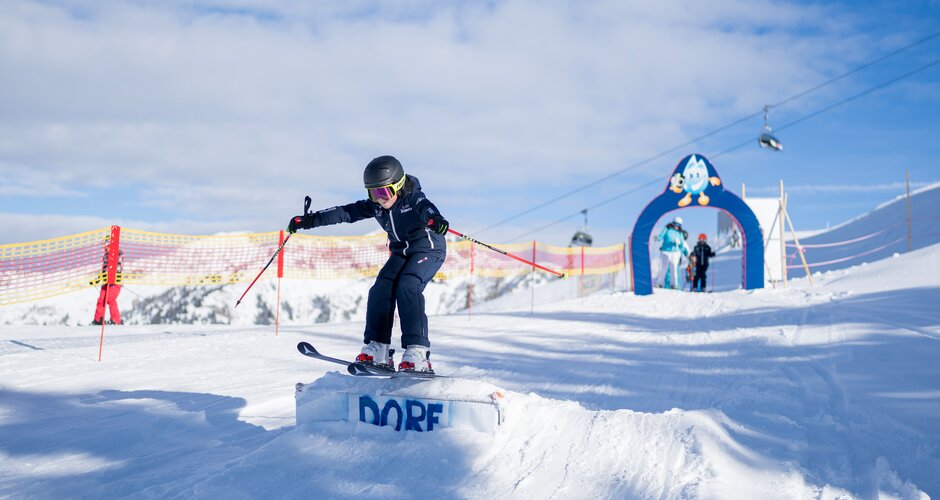 Kind in Skibekleidung springt über Schanze im Übungsbereich mit Skibogen und Sicherheitsnetz in Dorfgastein | © Dorfgasteiner Bergbahnen
