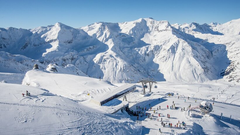 Bergstation einer Kabinenbahn von oben und daneben stehen Skifahrer und rundherum sind schneebedeckte Berge
