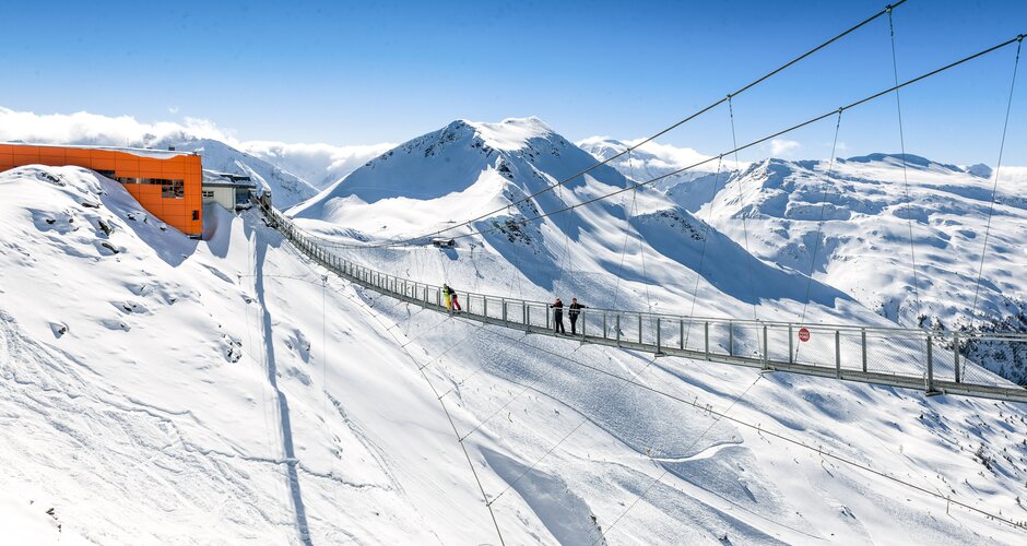 Hängebrücke spannt sich über einen Schneebedeckten Abgrund und an einem Ende ist eine orange Bergstation einer Kabinenbahn | © Gasteiner Bergbahnen AG, Wolkersdorfer