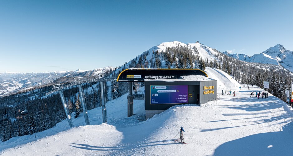 Skiers at Kaiblinggrat mountain station with snowy peaks and forest in the background. | © Hauser Kaibling | Josh Absenger