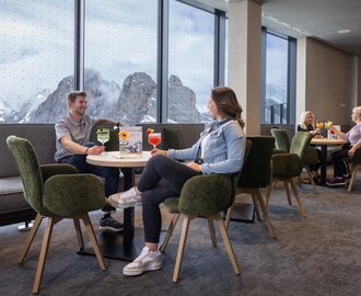 Menschen genießen Getränke an Tischen mit Blick auf verschneite Berge durch Panoramafenster. | © Harald Steiner