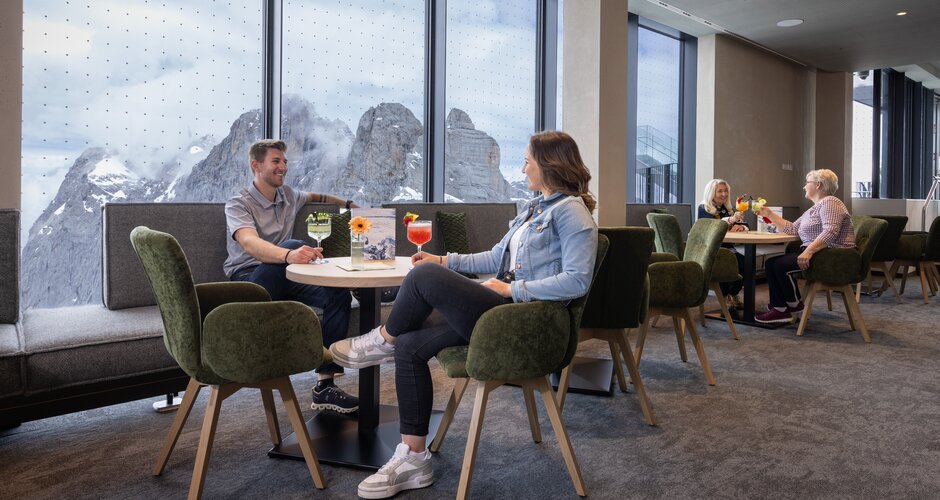 People enjoying drinks at tables with view of snow-covered mountains through panorama windows. | © Harald Steiner