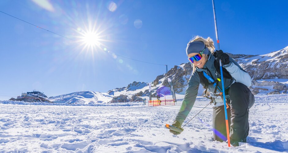 Woman kneeling in snow with probe and avalanche transceiver, sun and mountains in the background. | © René Eduard Perhab