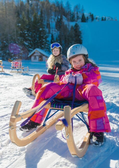 Smiling child in pink ski suit sledding in snow, wearing helmet, sunny day. | © Christine Höflehner