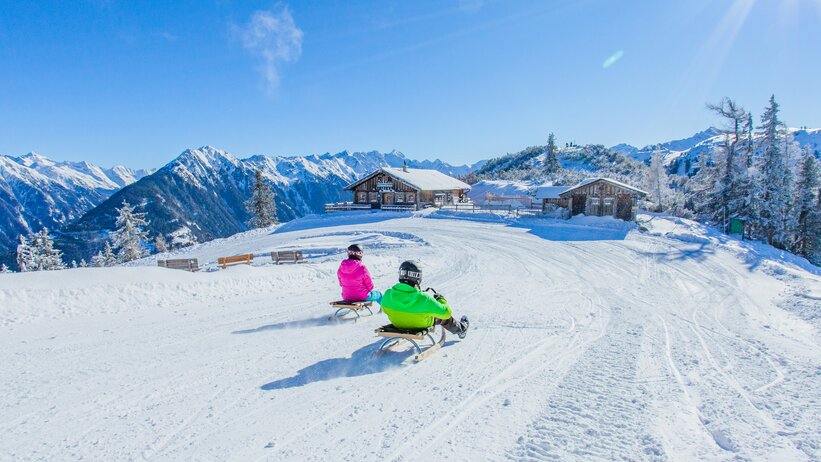 Two sledders ride towards alpine huts with snowy mountain backdrop. | © René Eduard Perhab