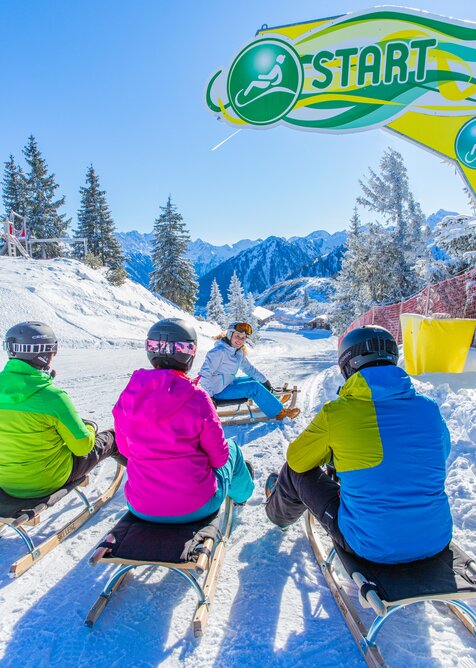 Sledders in colorful jackets at the start of the toboggan run with snowy mountains behind. | © René Eduard Perhab