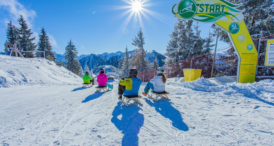 Vier Rodler starten am Hochwurzen-Rodelweg, sonniger Wintertag mit Bergen im Hintergrund. | © René Eduard Perhab